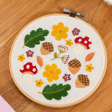 Load image into Gallery viewer, Finished Woodland Wonder embroidery hoop with red mushrooms, acorns, leaves and flowers on a wooden table