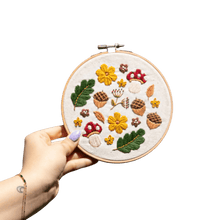 Load image into Gallery viewer, Hand holding a finished Woodland Wonder embroidery hoop with mushrooms, acorns, leaves and flowers on a plain background