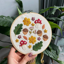 Load image into Gallery viewer, Hand holding a Woodland Wonder embroidery hoop with mushrooms, acorns and flowers in front of green houseplant leaves