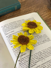Load image into Gallery viewer, Two finished yellow and brown beaded sunflowers with green stems lying on an open book
