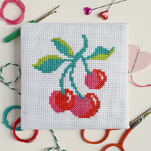 Load image into Gallery viewer, Close-up of the finished cherries cross stitch design on white Aida fabric with colourful threads and scissors
