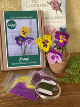 Load image into Gallery viewer, Pansy Beaded FloweHand holding yellow beaded pansy petals being made with wire from the Pansy Beaded Flower Craft Kitr Craft Kit box with purple and yellow seed beads, wire and finished beaded pansies
