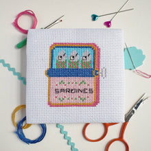 Load image into Gallery viewer, Close-up of a finished sardine tin cross stitch with the word Sardines, surrounded by threads and scissors
