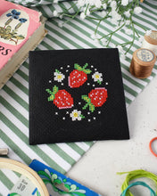 Load image into Gallery viewer, Finished strawberry patch mini cross stitch design on black Aida fabric with strawberries and daisies, surrounded by sewing supplies
